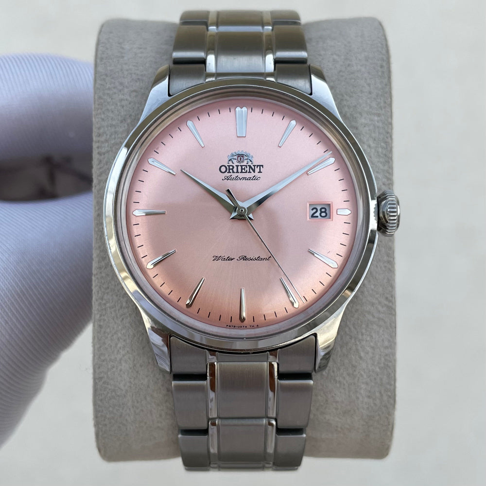 An Orient Water Standard wrist watch with a pink dial and a silver stainless steel case and bracelet, displayed on a gray velvet watch stand with a hand holding the left side.