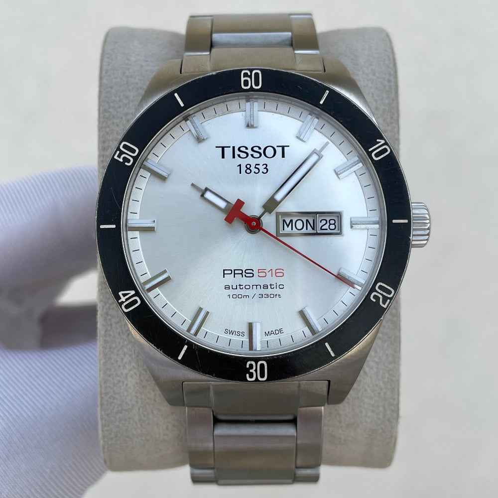 A Tissot PRS 516 automatic watch with a silver stainless steel case and bracelet, a white dial with a black rotating bezel, and a red seconds hand, displayed on a light gray stand.
