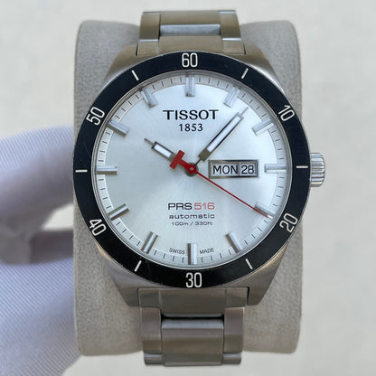 A Tissot PRS 516 automatic watch with a silver stainless steel case and bracelet, a white dial with a black rotating bezel, and a red seconds hand, displayed on a light gray stand.