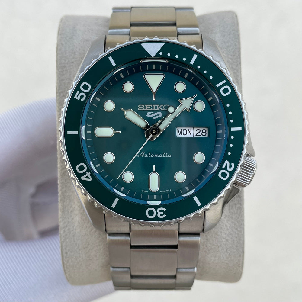 A Seiko dive watch with a dark green dial and bezel, featuring a stainless steel link bracelet and a day/date window, displayed on a light gray stand.