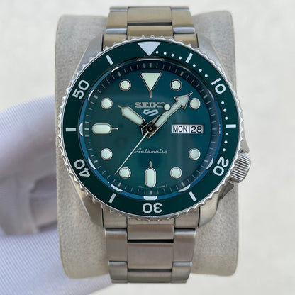 A Seiko dive watch with a dark green dial and bezel, featuring a stainless steel link bracelet and a day/date window, displayed on a light gray stand.
