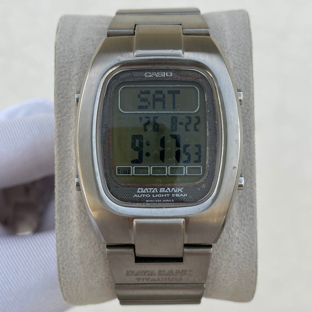 A Casio Data Bank titanium analog-digital watch with a brushed silver-tone metal case and bracelet, displayed on a light gray velvet wrist strap. The watch features a square digital LCD screen showing the time as 9:17 and the day as SAT.