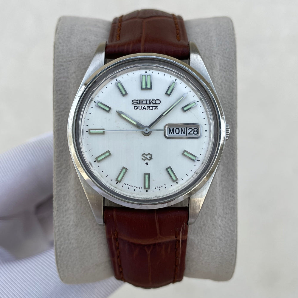 A vintage Seiko Quartz wristwatch with a white dial, silver-tone stainless steel case, and a brown leather strap, displayed on a gray cushion held by a hand.