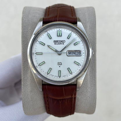A vintage Seiko Quartz wristwatch with a white dial, silver-tone stainless steel case, and a brown leather strap, displayed on a gray cushion held by a hand.