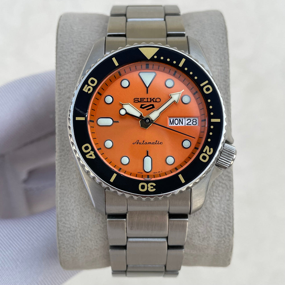 A Seiko dive watch with a bright orange dial, black rotating bezel, and stainless steel bracelet, displayed on a gray cushion.