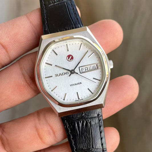 A hand holding a Rado watch with an octagonal silver-toned case, white dial, and black crocodile-embossed leather strap against a blurred gray background.