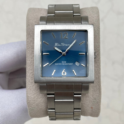 A rectangular stainless steel wristwatch with a blue dial and silver-tone metal bracelet, displayed on a beige cushion. The watch face shows the brand name 'Ben Shen'.